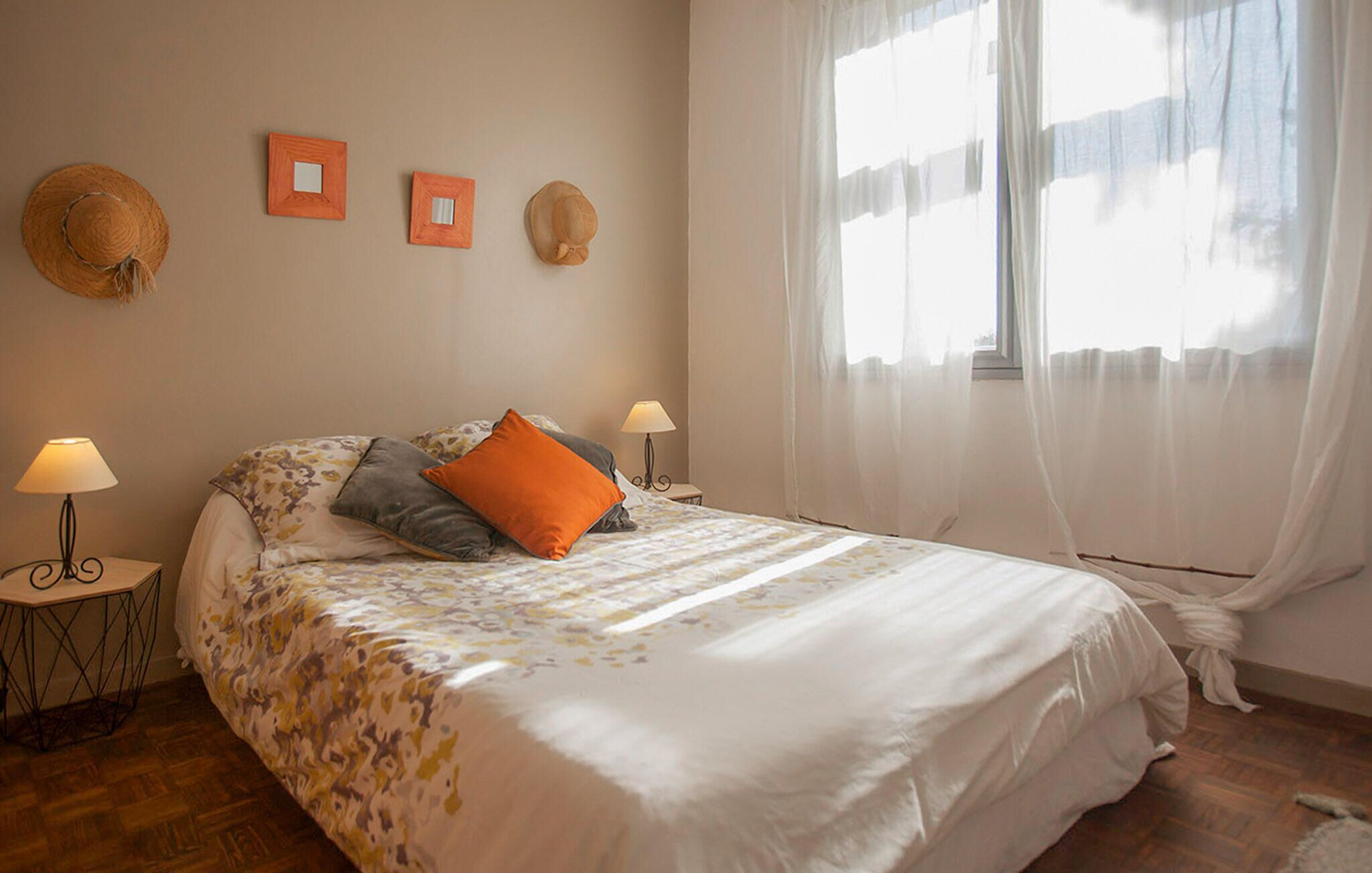 A bedroom with a bed, two nightstands, a window with sheer curtains, and wall-mounted hats.