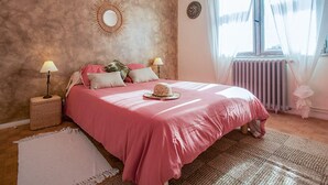 Een slaapkamer met een groot bed, twee bedlampen, een decoratieve spiegel en een raam met dunne gordijnen.
