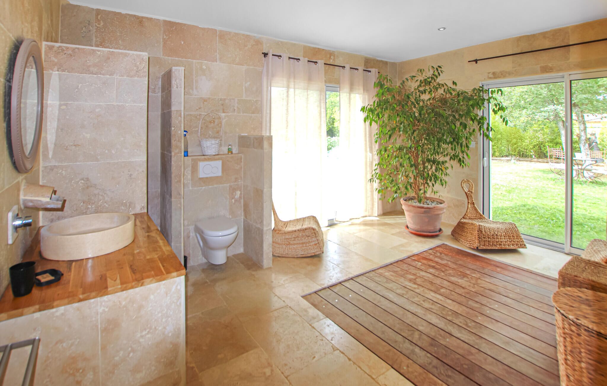 A bathroom with a sink, toilet, and shower. There is a large potted plant and wicker furniture near the window.