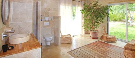 A bathroom with a sink, toilet, and shower. There is a large potted plant and wicker furniture near the window.