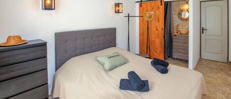 Une chambre à coucher comprenant un lit, une commode, une porte coulissante en bois, une salle de bain avec un lavabo et un miroir, et un luminaire fixé au mur.