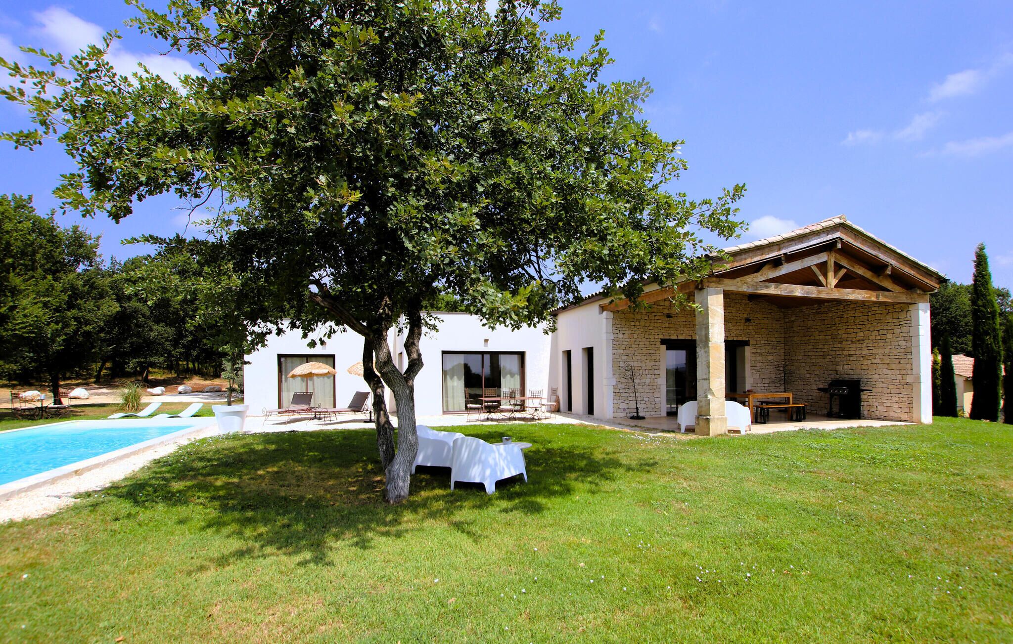 A modern house with a swimming pool, outdoor seating, and a large tree in the foreground.