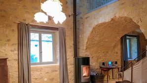 Una camera rustica con parete in pietra, stufa a legna, una finestra con tende e un tavolo con un computer.