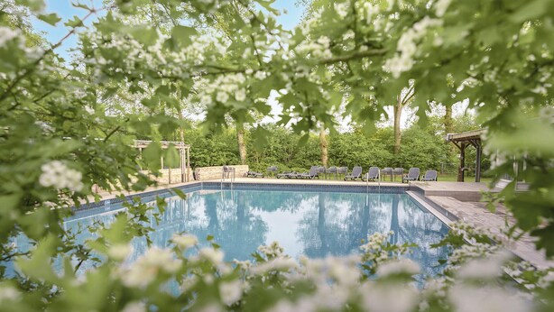 A rectangular swimming pool with a clear blue water surface, surrounded by a stone deck. There are several lounge chairs aligned along the pool's edge, and a wooden pergola structure is visible in the background. - Cozy home in Lemele with heated swimming pool (Lemele)