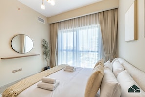 A hotel room with a bed, a bedside table, a mirror, a window with curtains, and a view of the city. - Chic 1BR | Next to Metro | Brand New | BMS (Dubai)