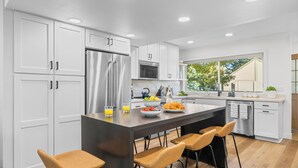 A modern kitchen with white cabinetry, a central island with bar stools, and a view of the outdoors. - Park City Getaway with Mountain Views, Hot Tub & Fire pit | High West Hideaway (Park City)