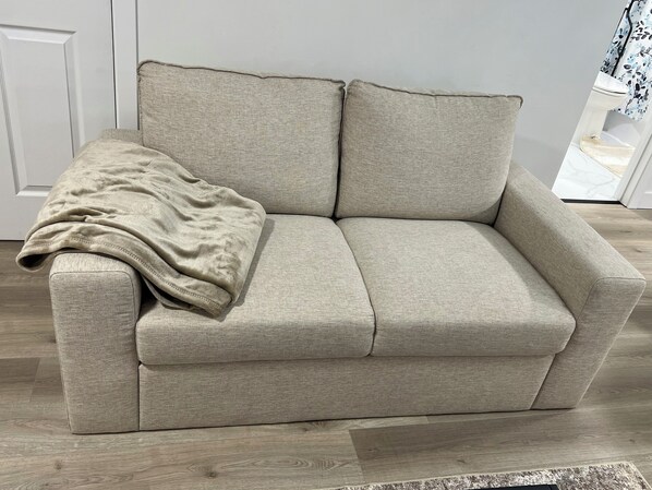 Un sofá color beige con una manta, ubicado en una habitación con piso de madera y una puerta blanca al fondo.