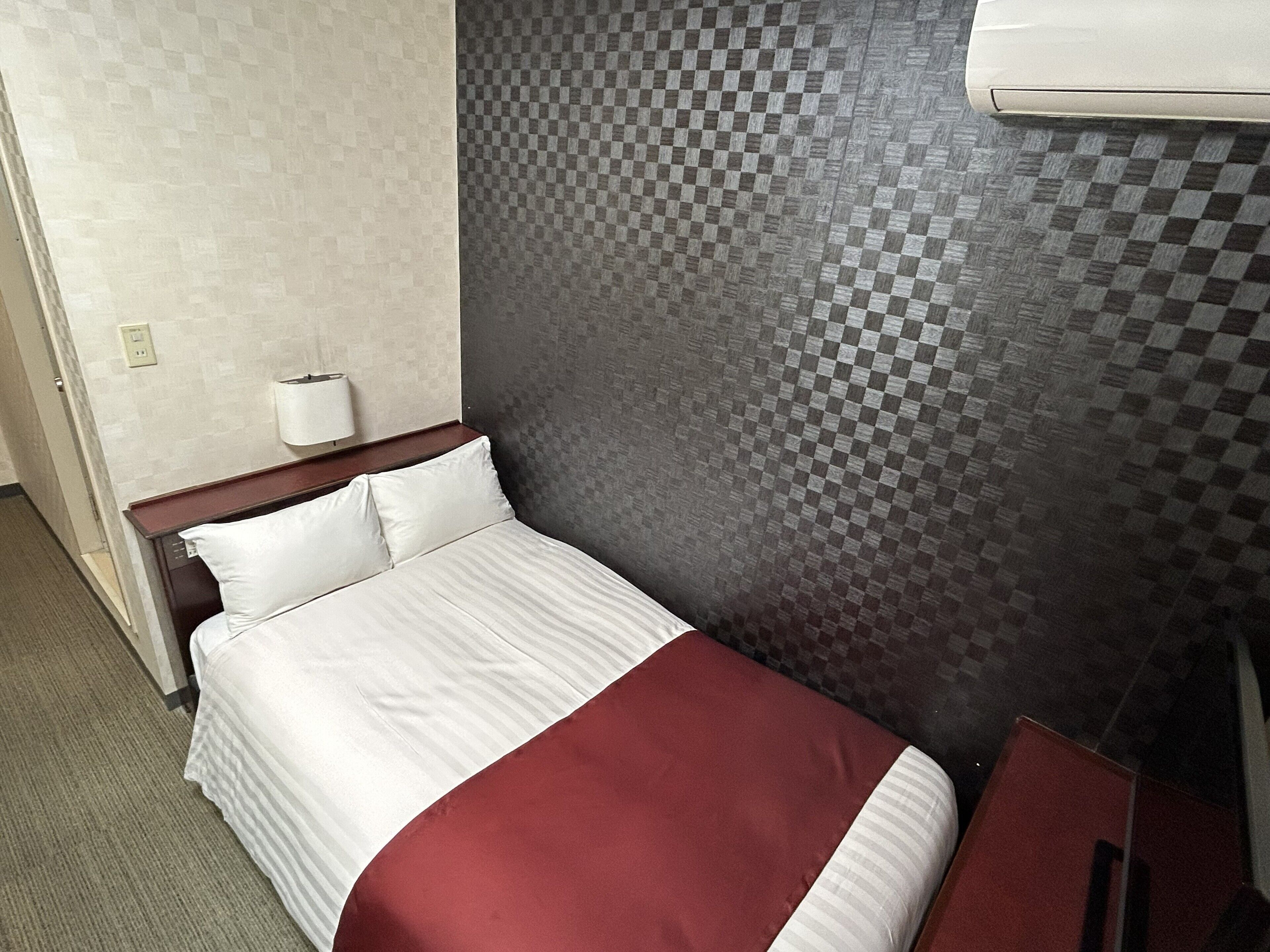 Ein Hotelzimmer mit einem Bett, einem Nachttisch aus Holz, einer Wand-Klimaanlage und einer strukturierten Wand.