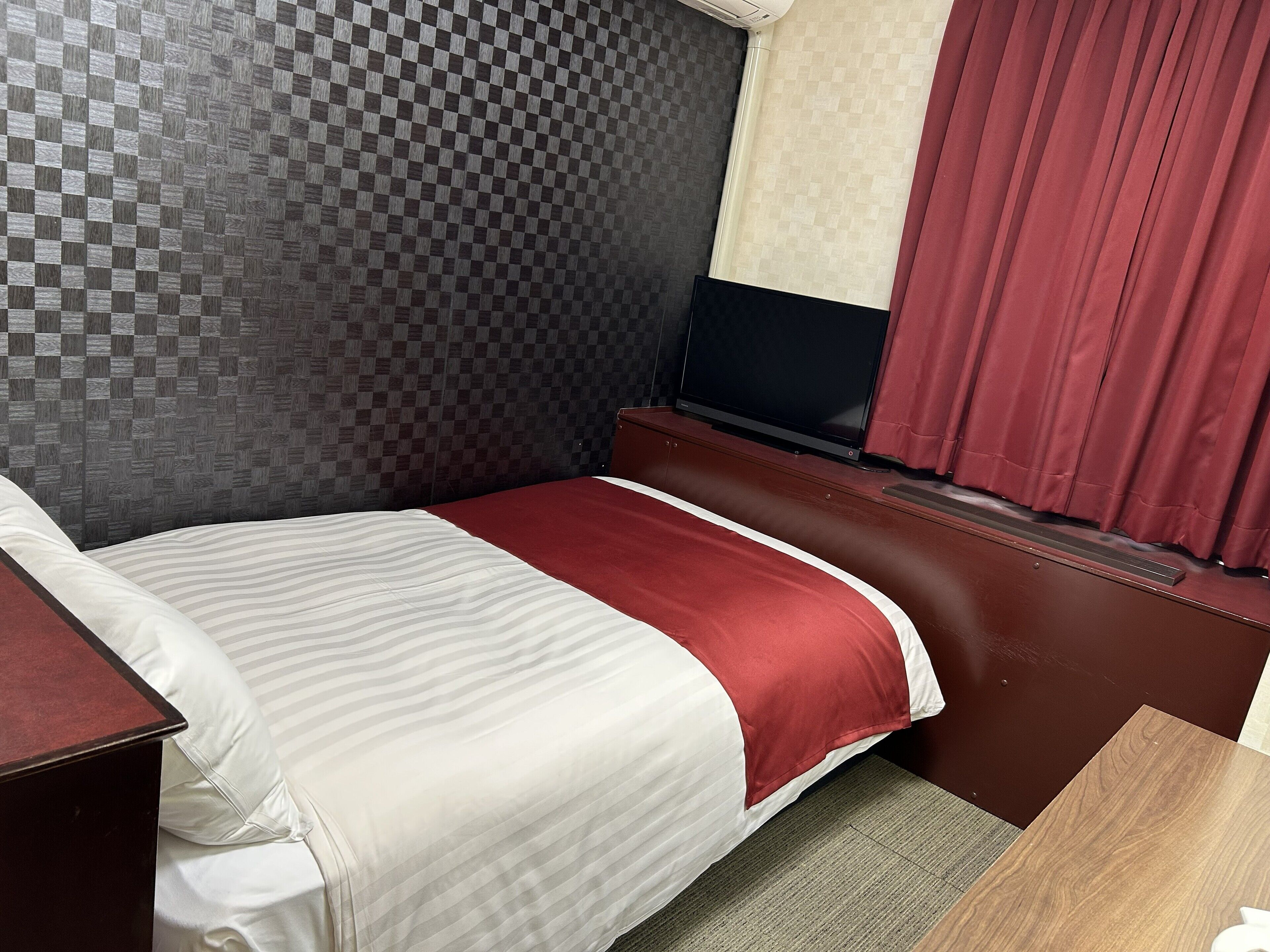 Ein Hotelzimmer mit kariertem Kopfteil, einem Bett mit rot-weißer Bettdecke, einem Fernseher auf einem Holztisch und einem roten Vorhang.