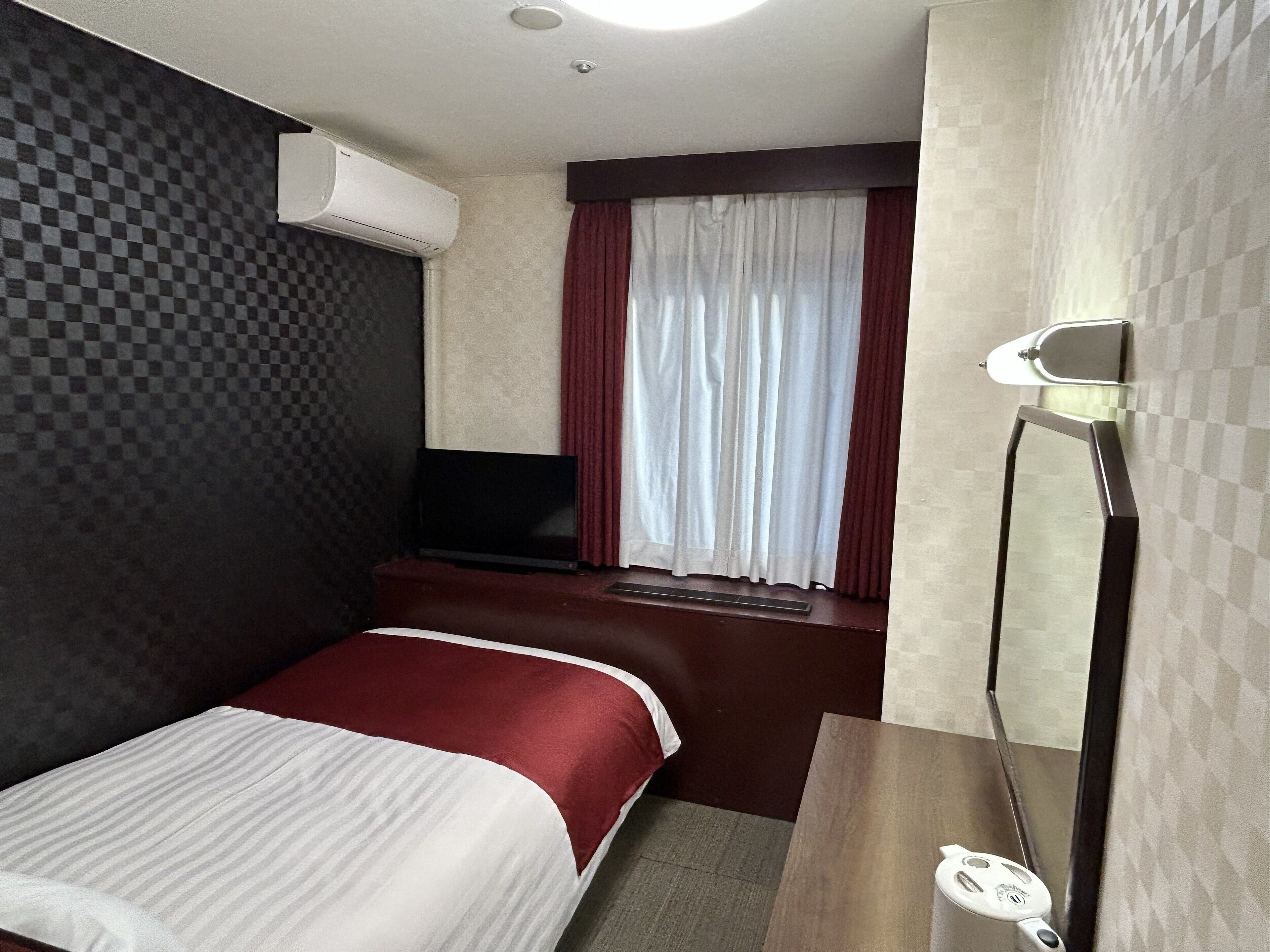Ein kleines Hotelzimmer mit Bett, Fernseher, Fenster mit Vorhängen und Spiegel.