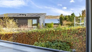 Una casa moderna con un ventanal grande, vistas a un lago y un seto en primer plano.