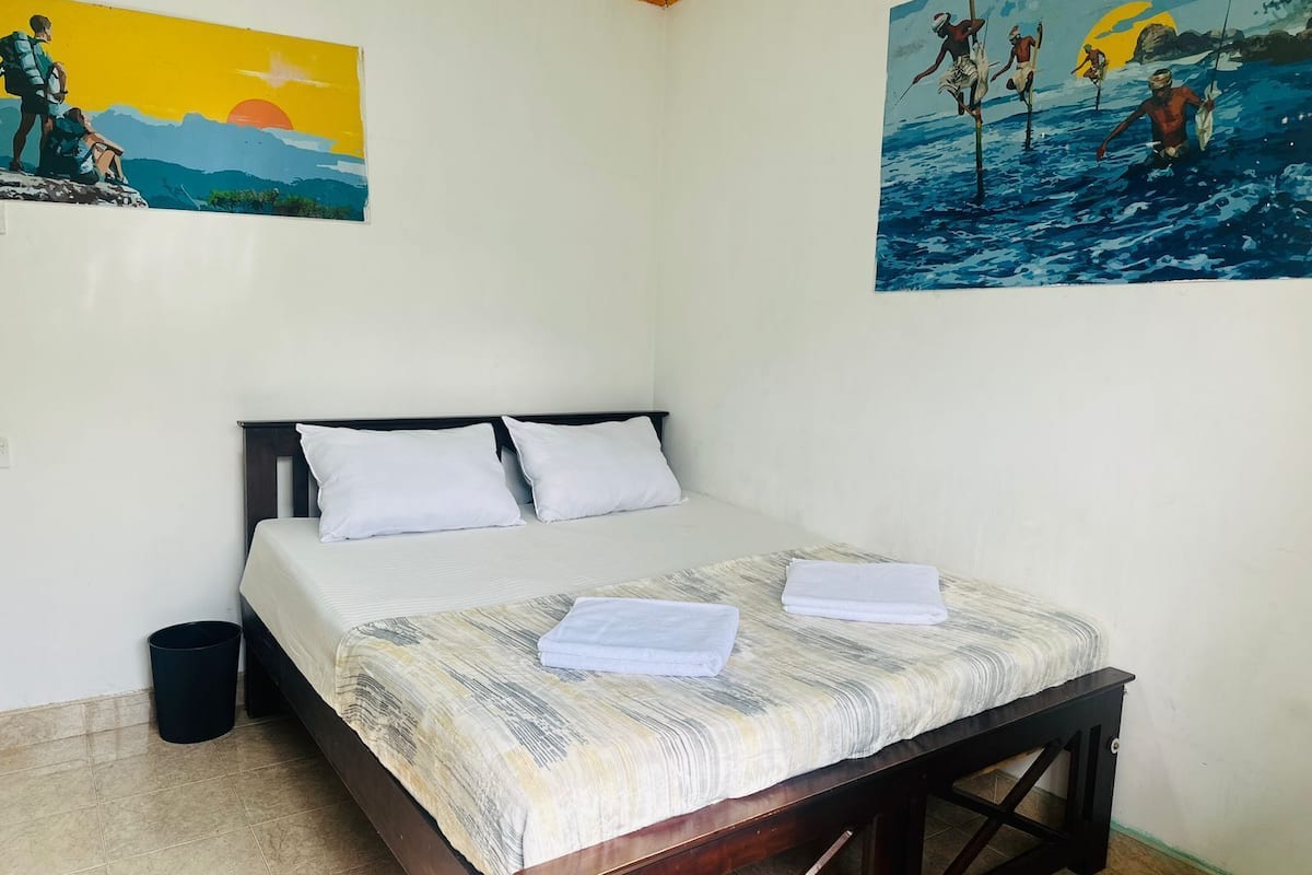A bedroom with a bed, pillows, a nightstand, a trash bin, and two paintings on the walls.