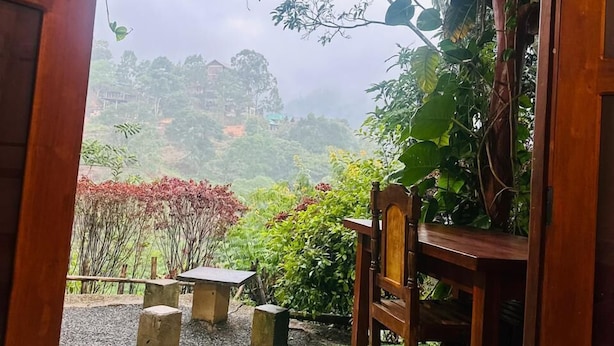 A wooden chair and table under a roofed area with a view of a lush garden and distant buildings. - Ella Hide Forest Cottage (Ella)