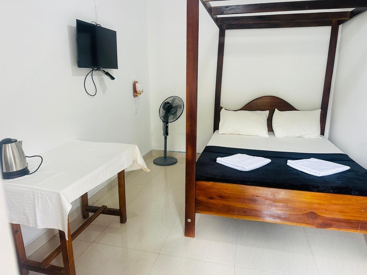 A room with a bed, a table, a kettle, a TV, and a fan.