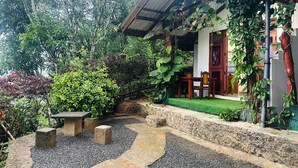 A covered outdoor seating area with stone benches, surrounded by a garden with various plants and a pathway leading to a building with a red door. - Ella Hide Forest Cottage (Ella)