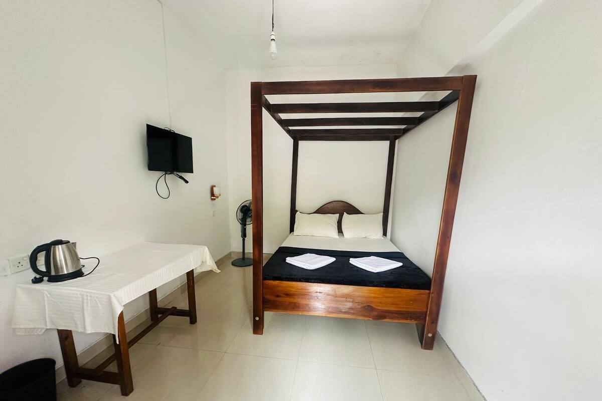 A small, clean room with a wooden bed, a white table with a kettle, and a wall-mounted TV.