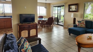 A living room with a sofa, a television, a dining area with chairs, a kitchen area with cabinets, and a balcony with a view.