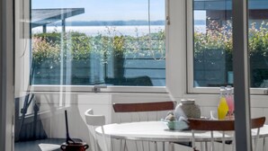 A room with a large window, a table, chairs, and a view of a body of water and greenery. - "Hjalma" - 250m to the fjord by Interhome (Farsø)