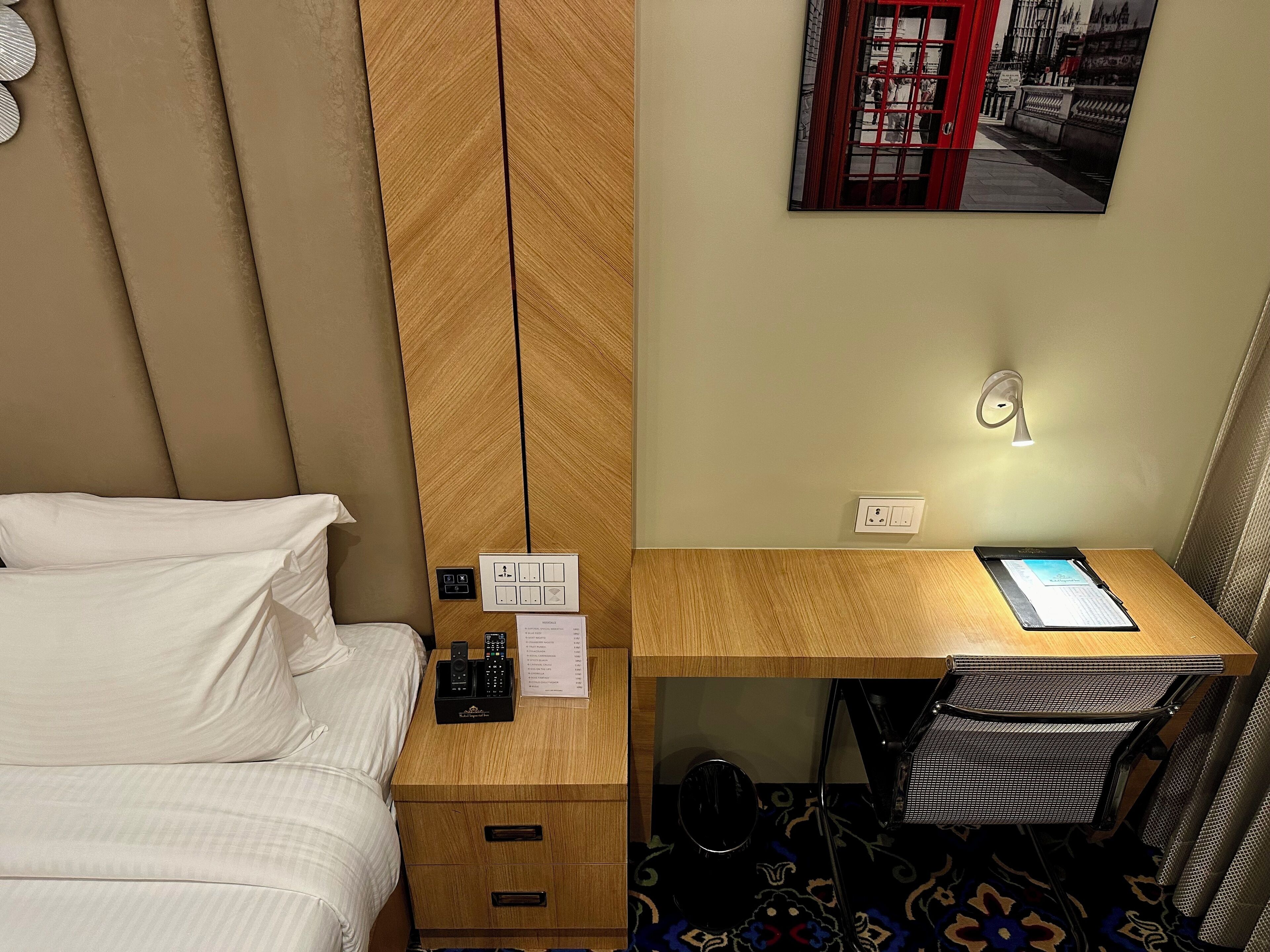 A hotel room with a bed, a wooden nightstand with a remote control, a desk with a chair, and a wall-mounted light.
