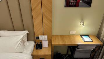 A hotel room with a bed, a wooden nightstand with a remote control, a desk with a chair, and a wall-mounted light.