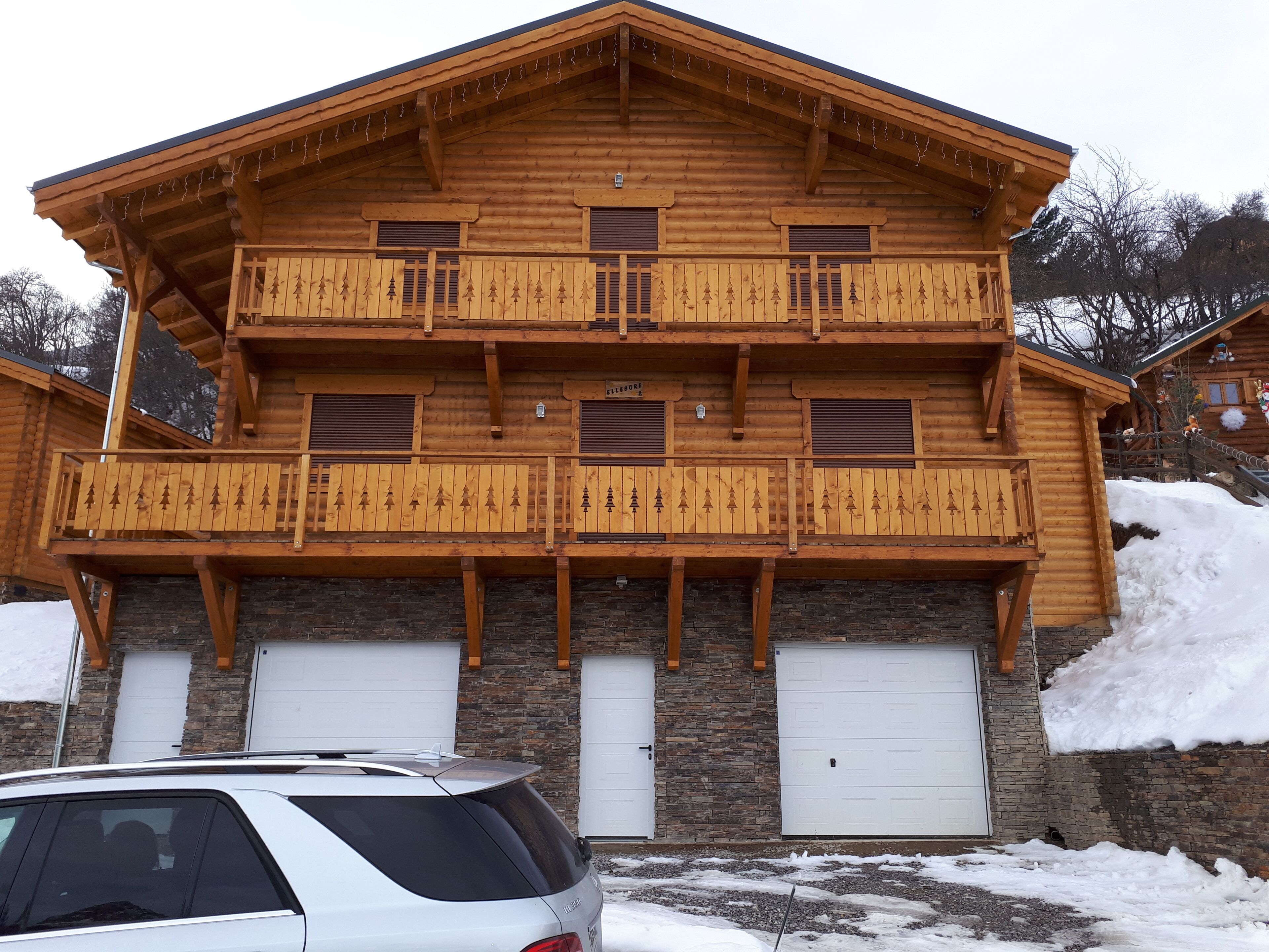 Eine zweistöckige Blockhütte mit Balkon, Garage und verschneitem Gelände.