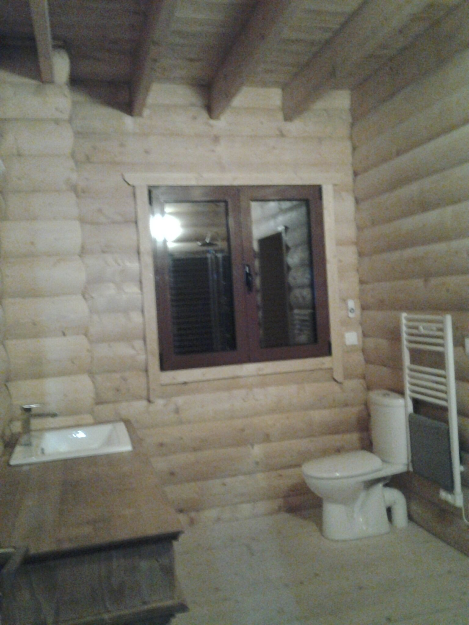 Une salle de bain rustique, aménagée dans une cabane en rondins, avec un lavabo en bois, des toilettes et une fenêtre qui s’ouvre comme une porte.