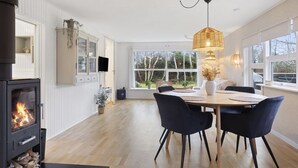 Una cocina moderna con estufa de leña, comedor con mesa redonda y sillas, y amplias ventanas con vistas al exterior.