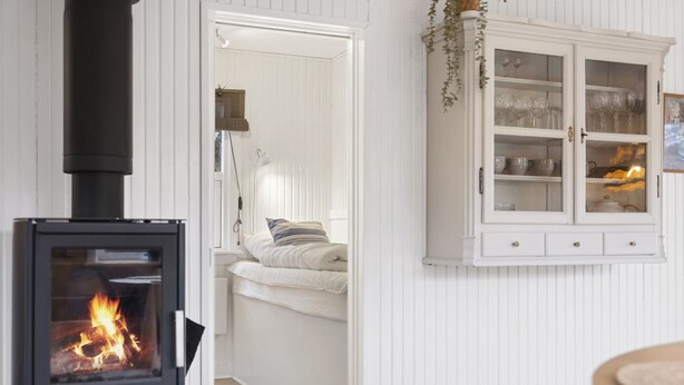 Una habitación acogedora con una estufa de leña, un armario blanco con puertas de cristal y una cama con un almohadón a rayas.