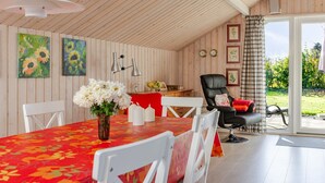 Et spiseområde med træloft, et bord med rødt blomstret dug, hvide stole, en vase med hvide blomster og en sort stol med ternet pude.