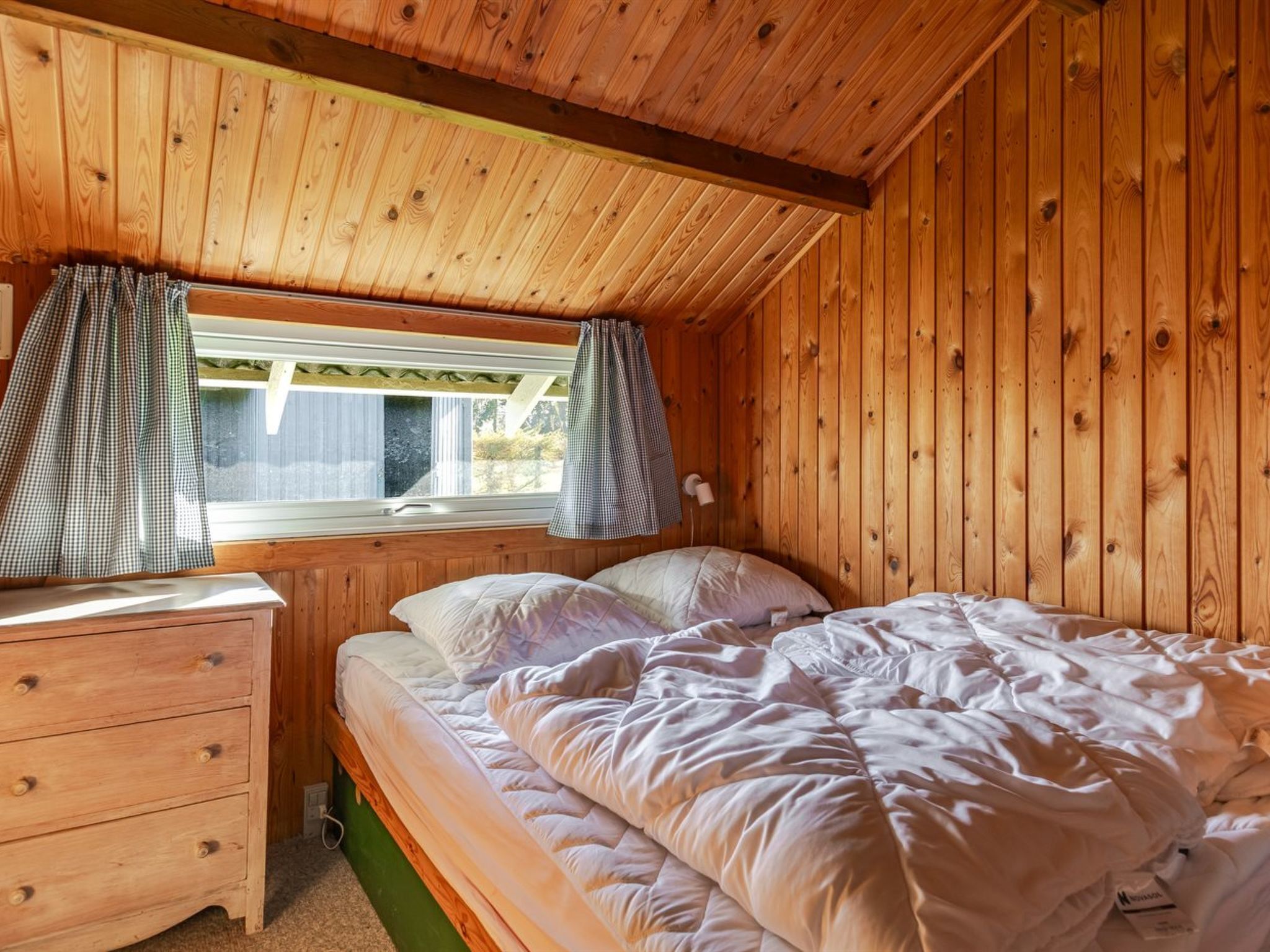 A wooden cabin bedroom with a single bed, a dresser, and a window with curtains.