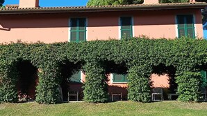 Un edificio con fachada rosa, contraventanas verdes y un seto cuidado frente a él.