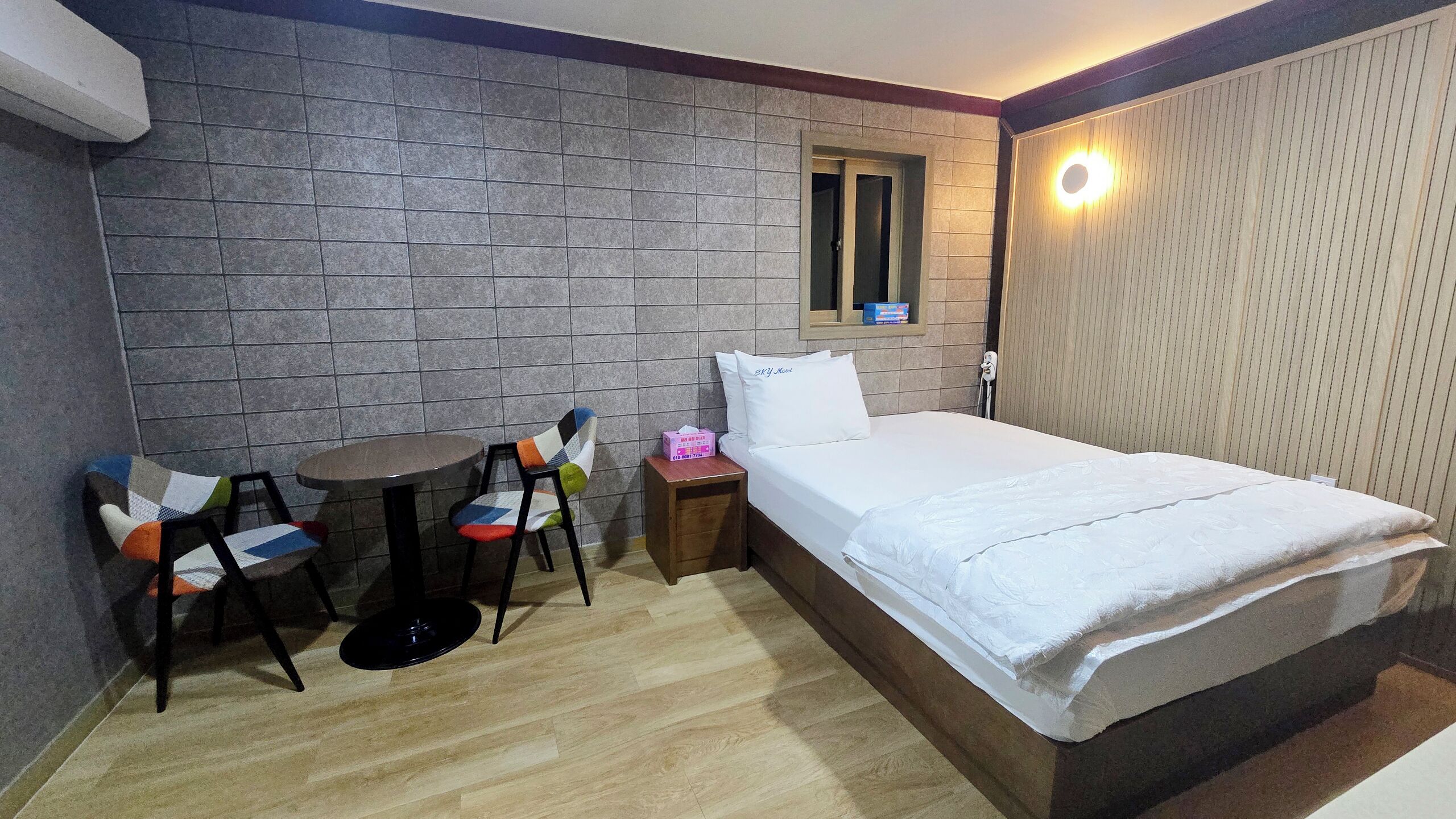 A hotel room with a bed, a small table, two chairs, a bedside table with a tissue box, and a wall-mounted light.