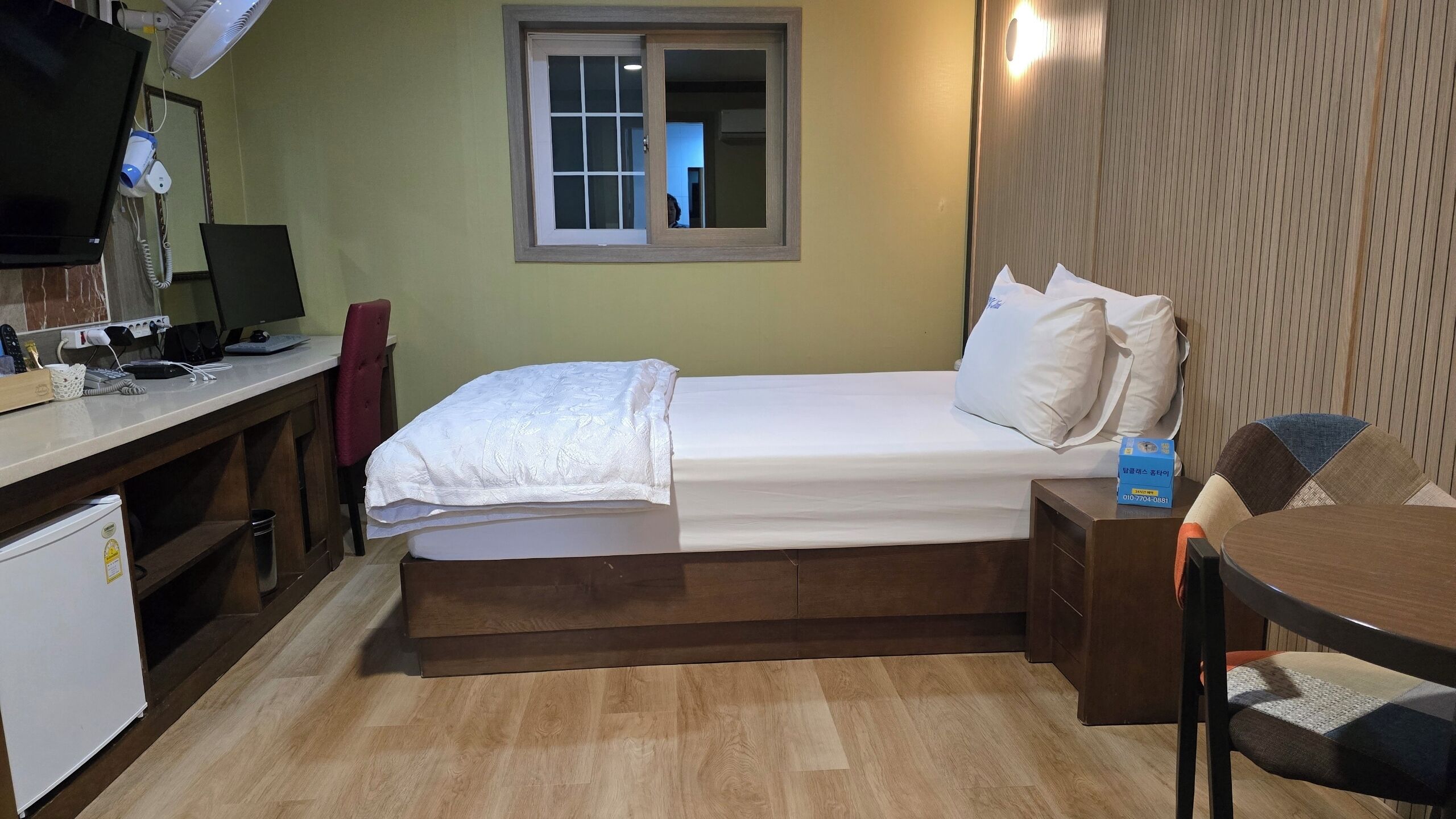 A compact hotel room with a single bed, a desk with a computer, a chair, a small table, and a window with a view.