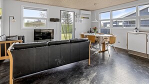 Ett modernt vardagsrum med en svart lädersoffa, ett matbord med stolar, en köksdel med skåp och stora fönster som släpper in naturligt ljus.