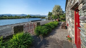 A stone building with a red door, a gravel path, and a metal fence leading to a body of water. - Pier Cottage Sneem (Sneem)