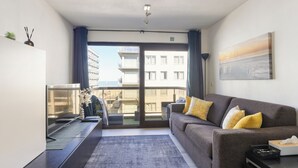 Un salon moderne avec un canapé, un téléviseur à écran plat, une table basse et une vue sur la ville grâce à de grandes fenêtres.