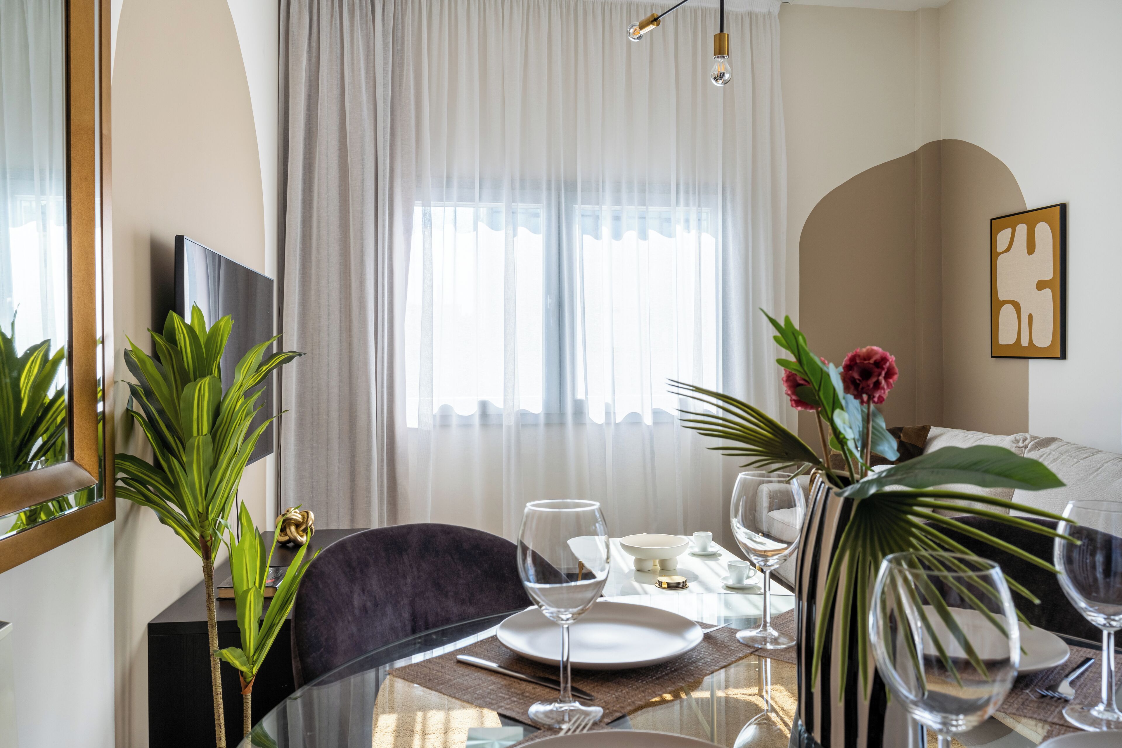 A dining area with a table set for four, a television on the wall, and a large window with curtains.