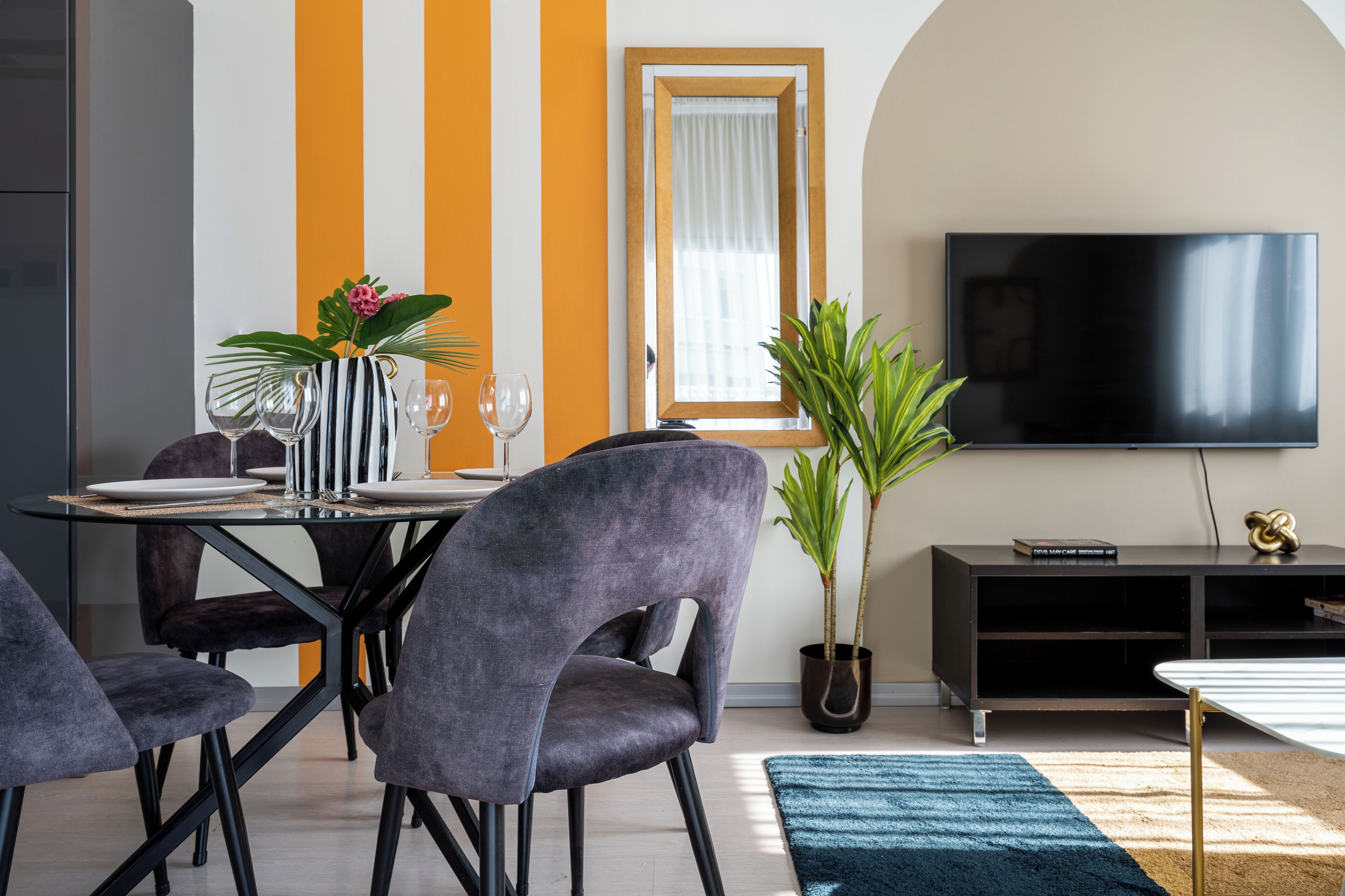 A dining area with a table set for four, surrounded by chairs, a TV mounted on the wall, and potted plants.