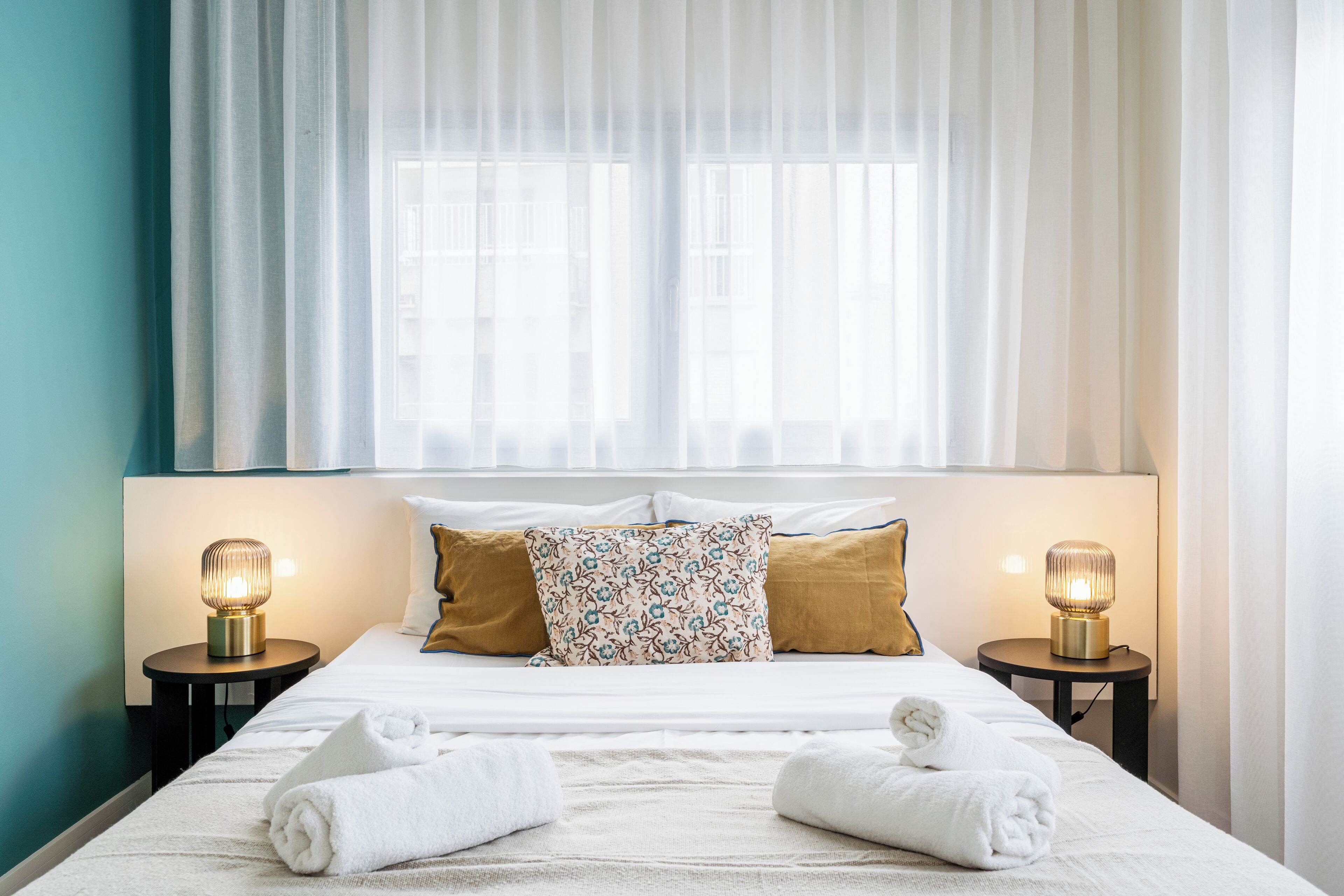 Een hotelkamer met een bed, twee bedlampen, een raam met dunne gordijnen en uitzicht op gebouwen.