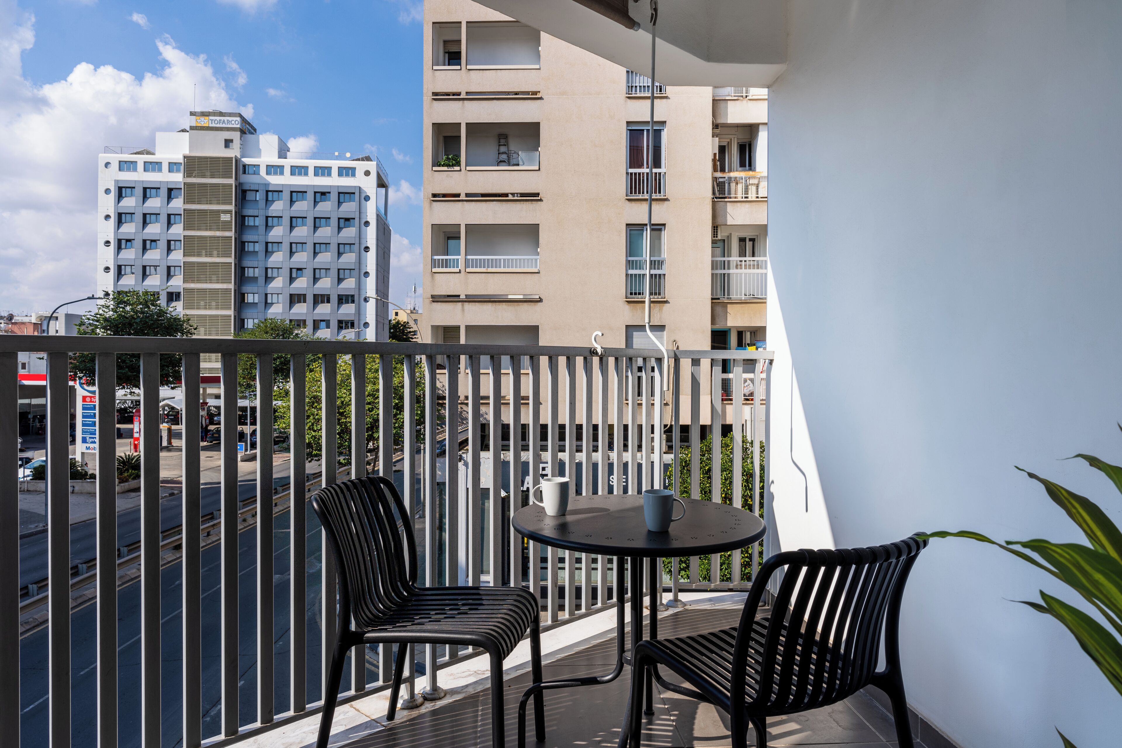 A balcony with two black chairs and a small table, overlooking a street with buildings and a gas station.
