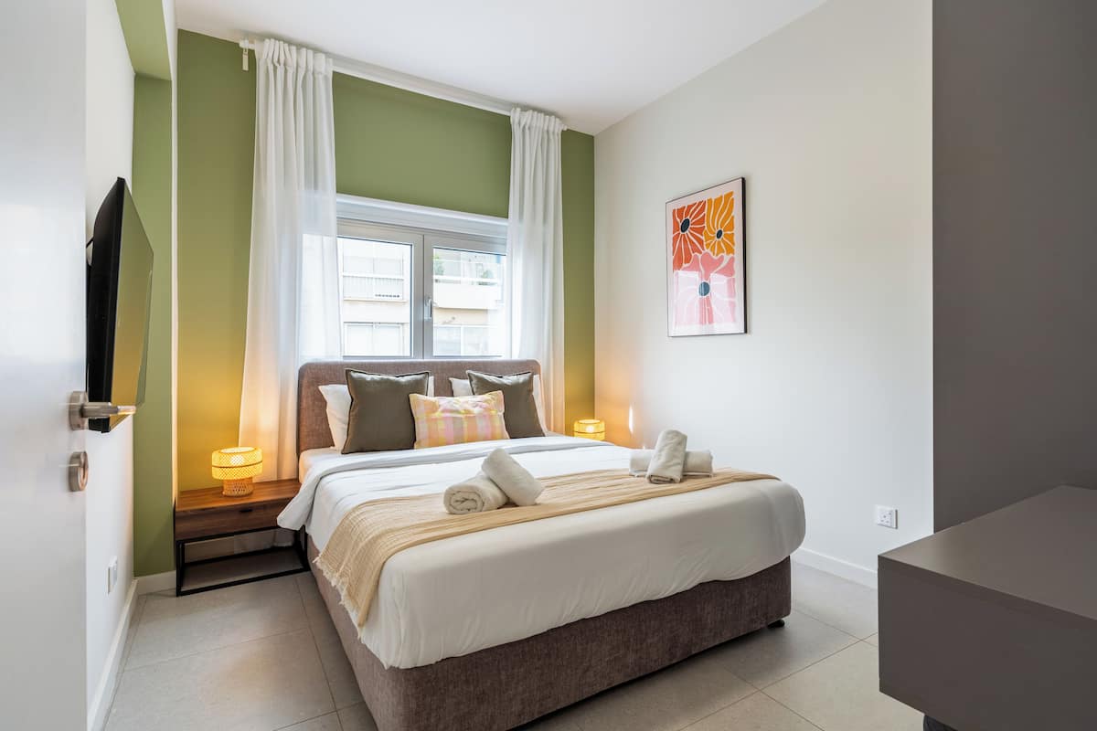 A bedroom with a bed, bedside lamps, a window with curtains, a wall-mounted TV, and a painting.