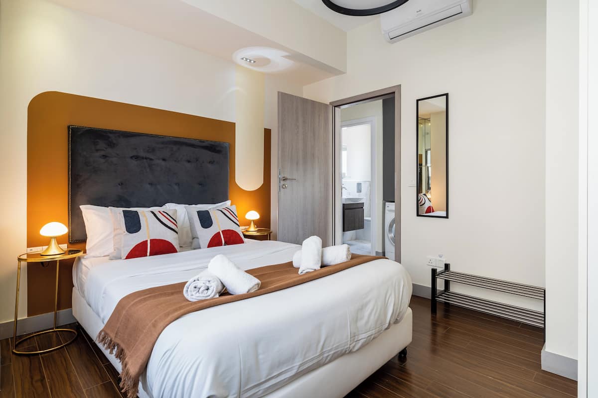 A modern hotel room with a large bed, bedside lamps, a wooden headboard, and a view of the bathroom through an open door.