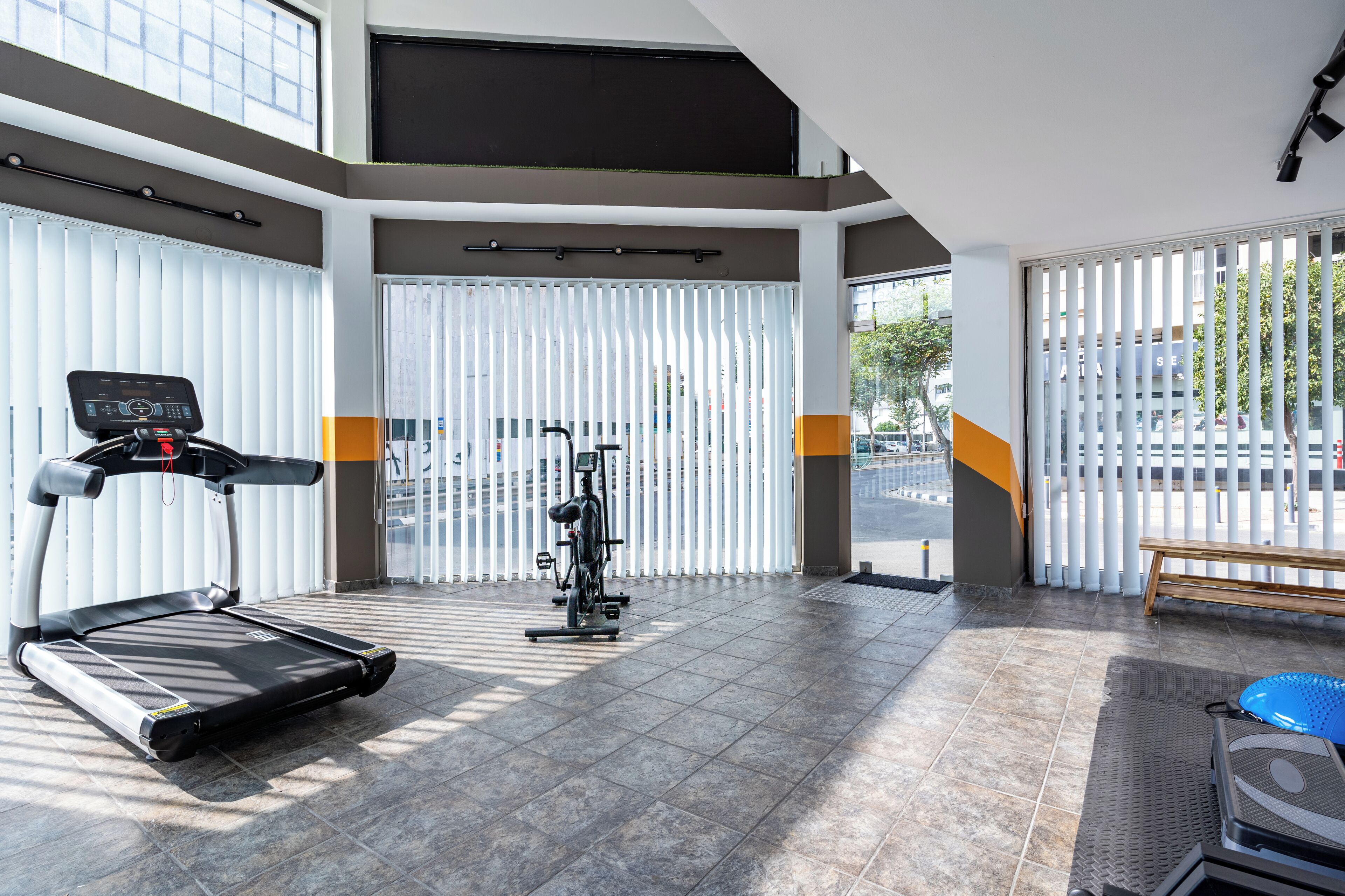 Ein Fitnessstudio mit Laufband, Crosstrainer und Gewichten, großen Fenstern und einer Sitzbank.