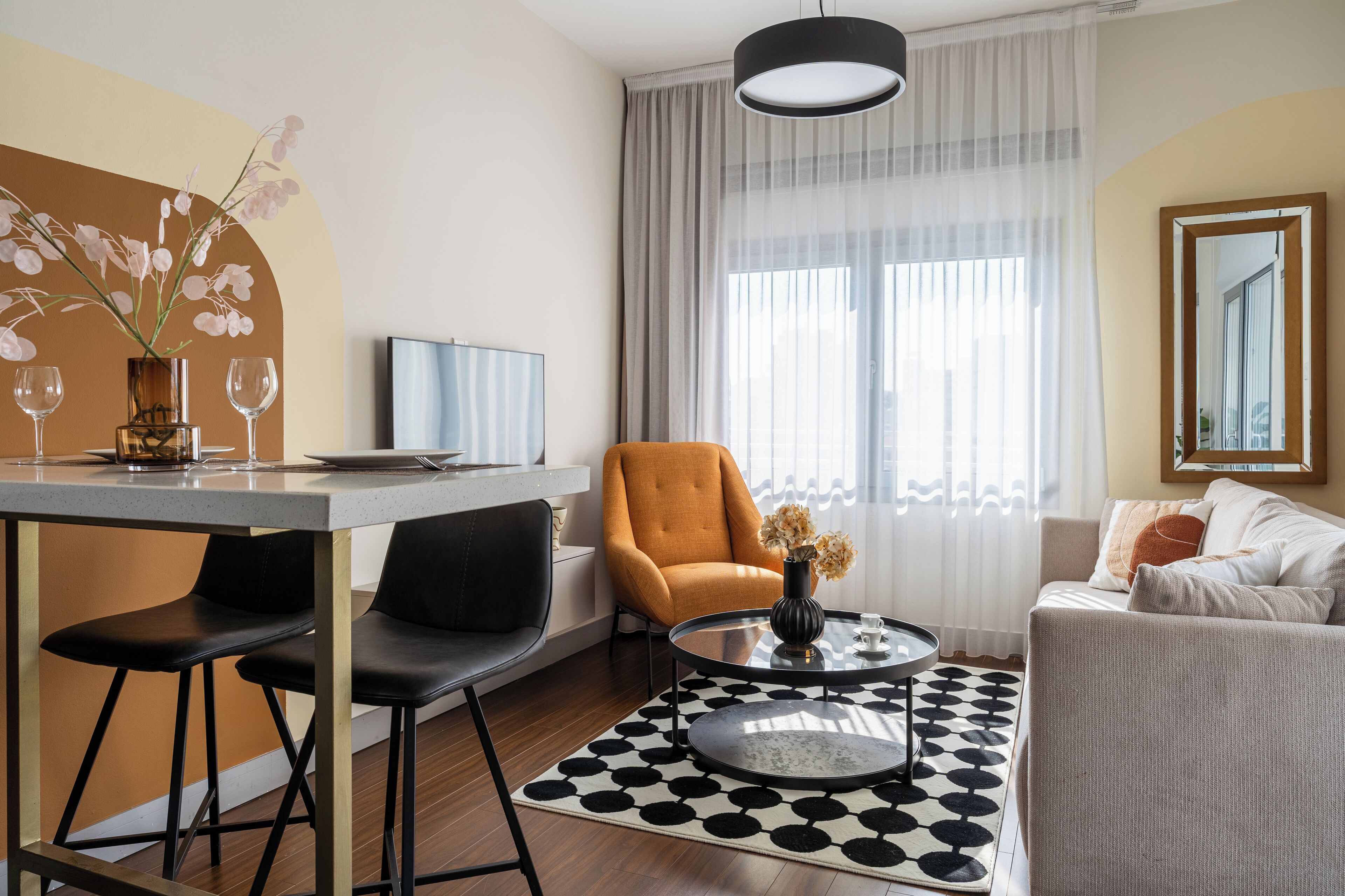 Una sala de estar moderna con comedor, un sofá, una mesa de centro y una silla. Hay un televisor en la pared y una ventana con cortinas.