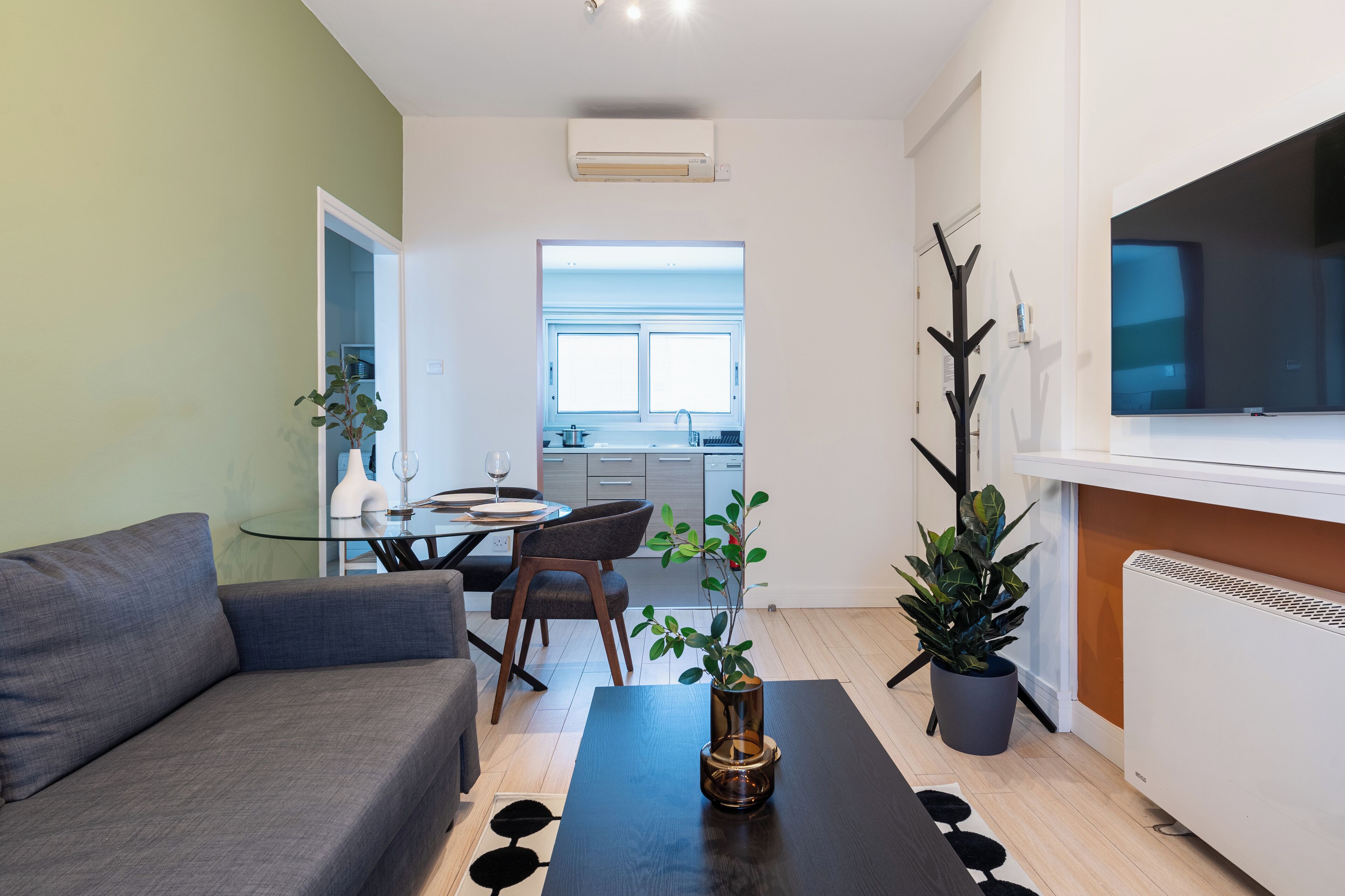 A modern living room with a sofa, a dining table with chairs, a coffee table with a vase, and a TV on the wall.