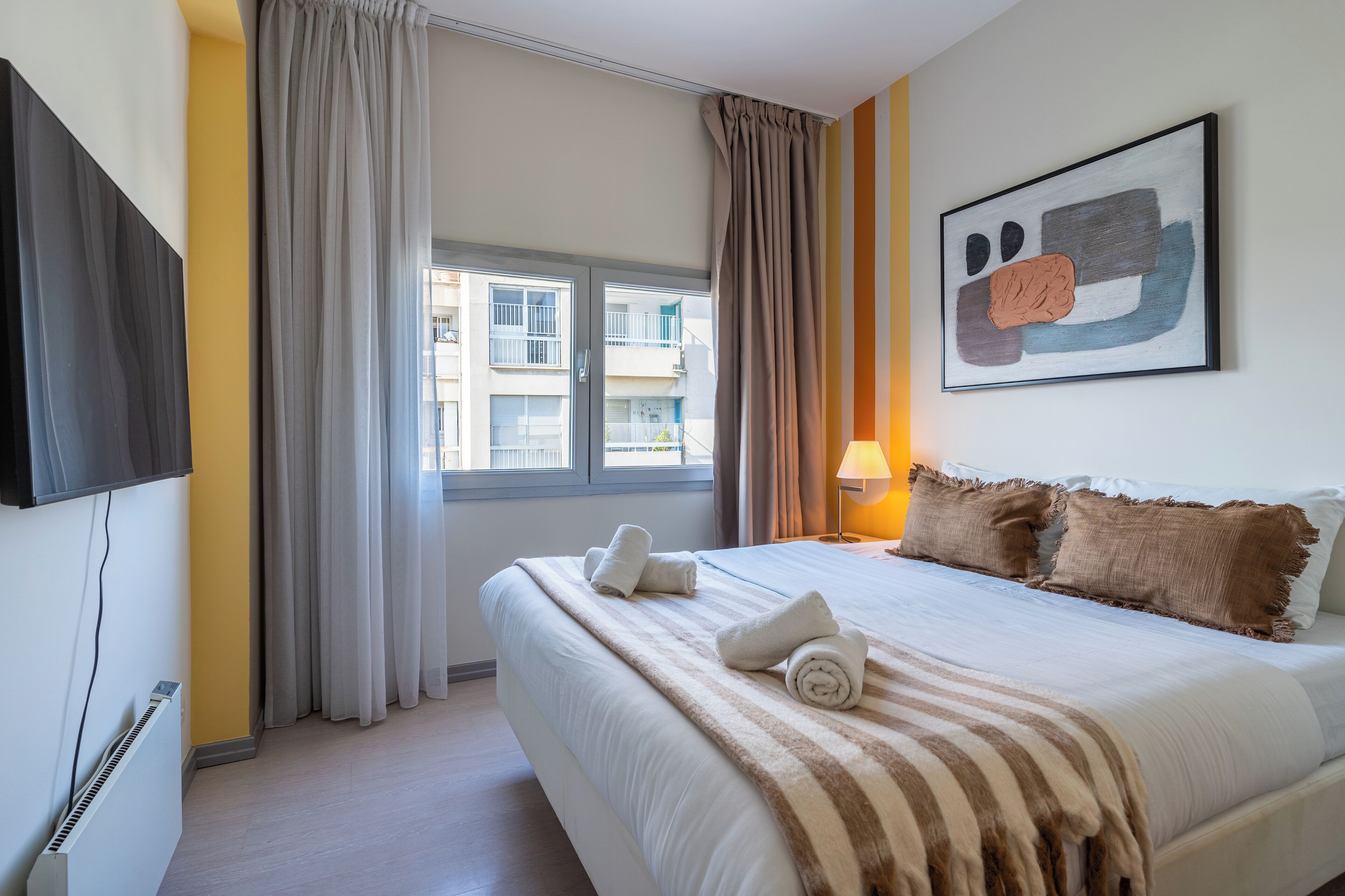 Une chambre d’hôtel avec un lit, une fenêtre avec des rideaux, un téléviseur fixé au mur et un tableau.