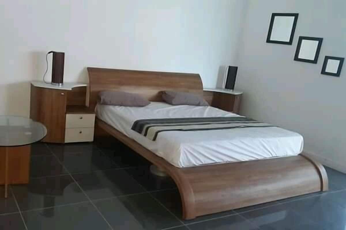 Un dormitorio moderno con una cama de madera, mesitas de noche, una mesa de vidrio y un estante fijado a la pared.