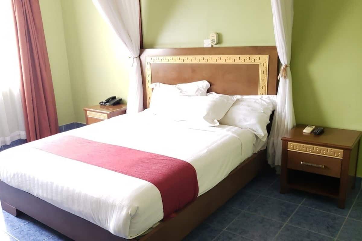 Una cama bien tendida con cabecera de madera, ropa de cama blanca y un detalle rojo. Hay dos mesitas de noche, una con un teléfono y la otra con un control remoto.