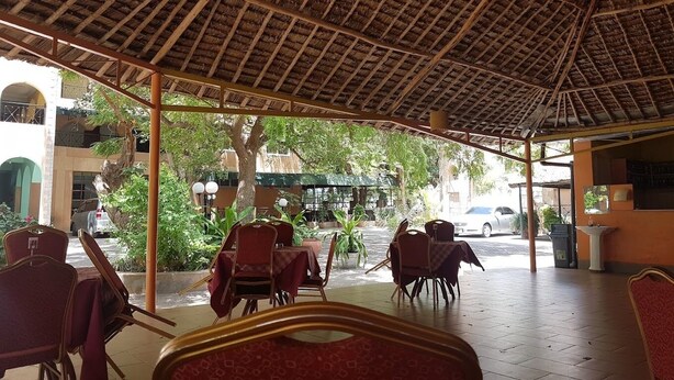Un café en plein air avec un toit de chaume, des chaises et des tables rouges, entouré de verdure et de bâtiments.