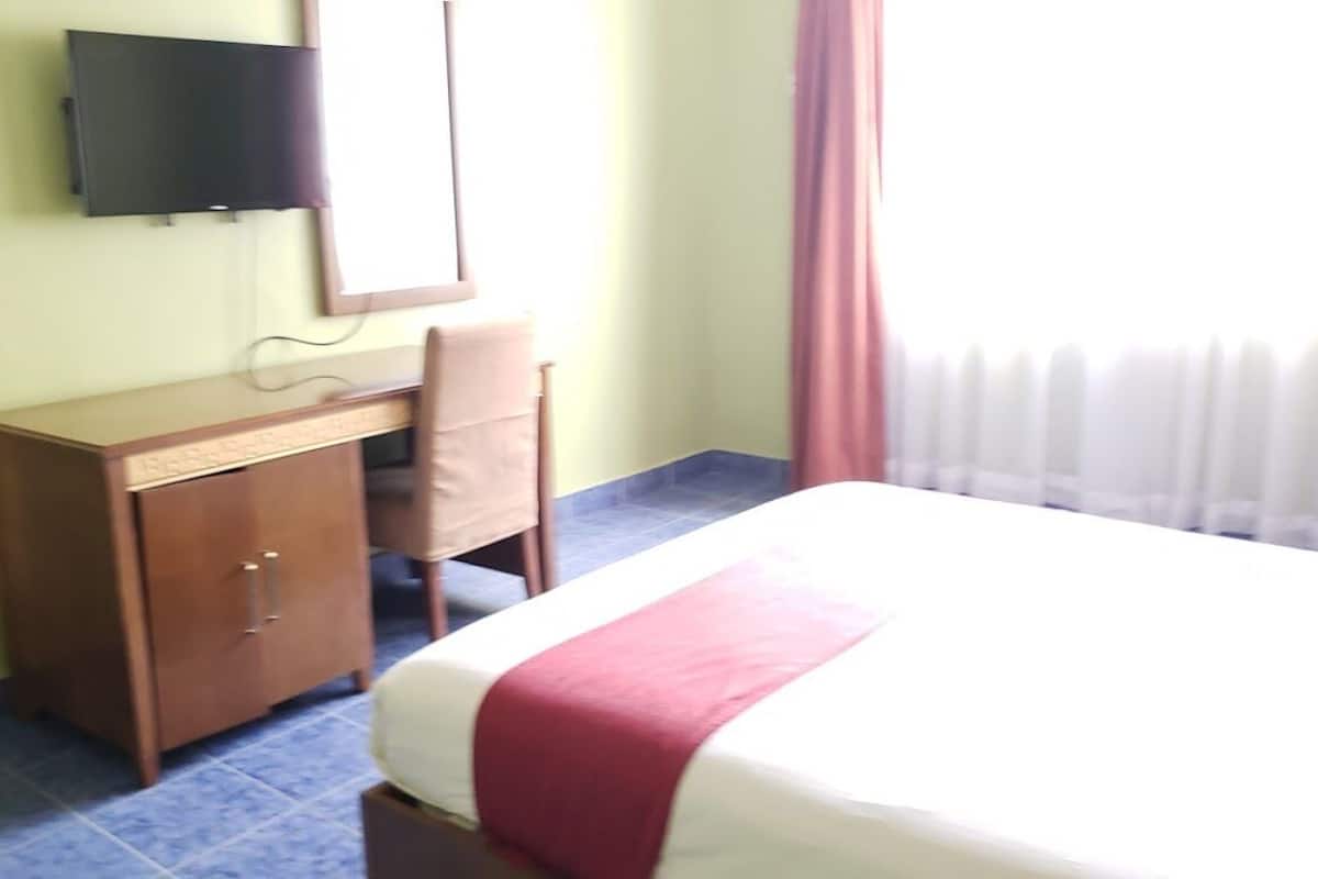 Habitación de hotel con cama, escritorio, silla, televisor montado en la pared y ventana con cortinas.